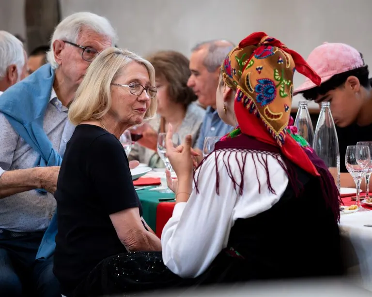 Mehrere Menschen sitzen an einem Tisch, eine Frau trägt traditionelle Kleidung und ein buntes Kopftuch, sie unterhalten sich.