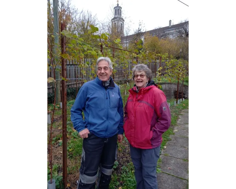 Zwei Personen stehen in einem herbstlichen Garten, lächeln in die Kamera, im Hintergrund ist ein Kirchturm zu sehen.