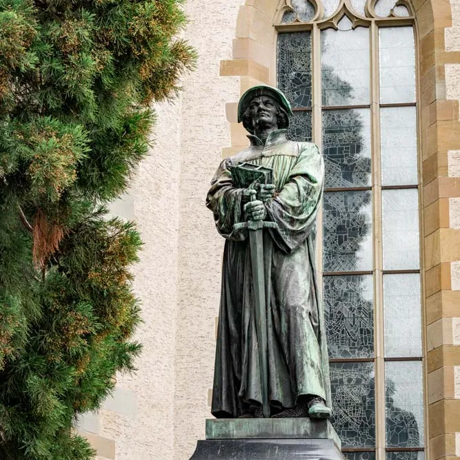 Bronzestatue von Zwingli mit Buch vor der Wasserkirche und Baum im Hintergrund.