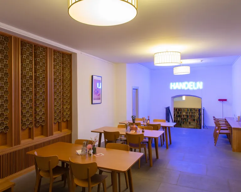 Heller Gastraum mit Holztischen und -stühlen, Weinregalen an der Wand und moderner Beleuchtung.