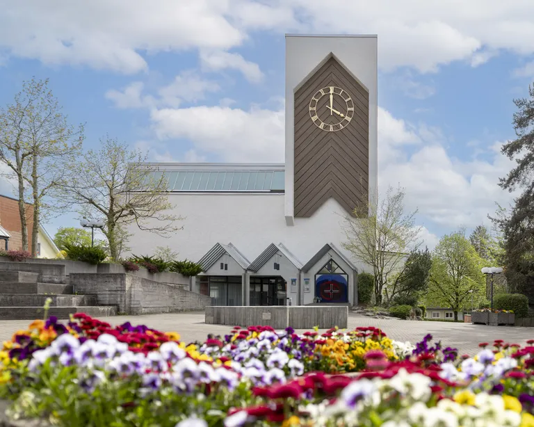 Kirche Oberengstringen mit modernem Turm und grosser Uhr, umgeben von kahlen Bäumen und Treppen an einem sonnigen Tag.