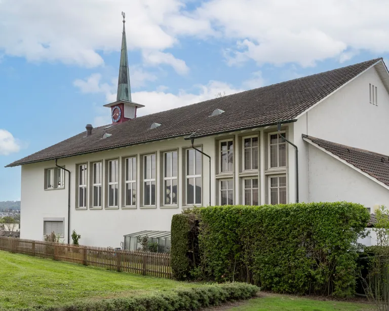 Kirchgemeindehaus Oberengstringen mit spitzem Kirchturm, grossen Fenstern und grünem Rasen im Vordergrund bei blauem Himmel.