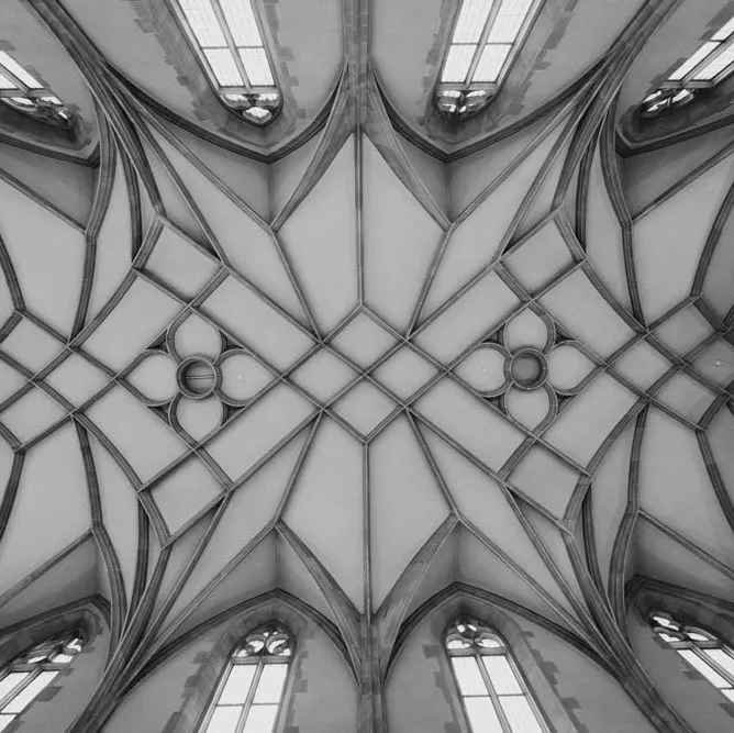 Blick nach oben auf ein kunstvoll gemustertes Gewölbe mit gotischen Fenstern in einer Kirche, schwarz-weiß aufgenommen.