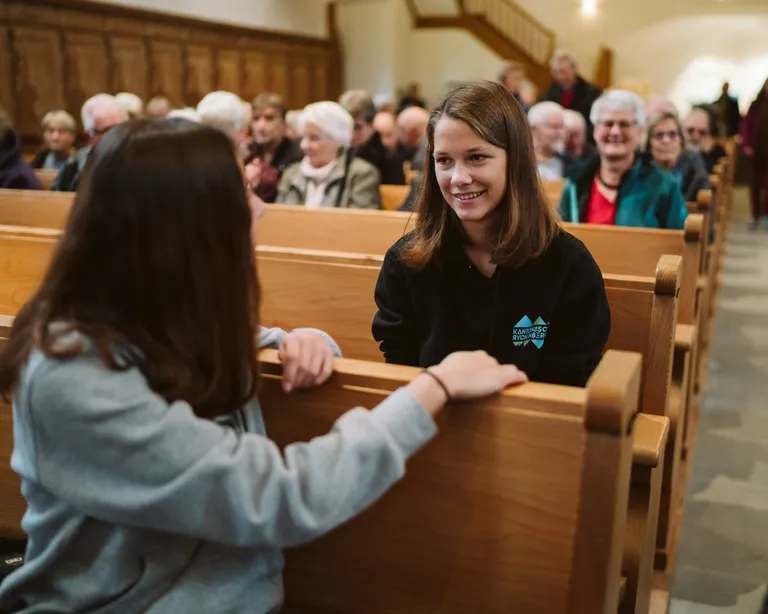Zwei junge Frauen unterhalten sich lächelnd in einer Kirchenbank, hinter ihnen sitzen viele ältere Menschen.