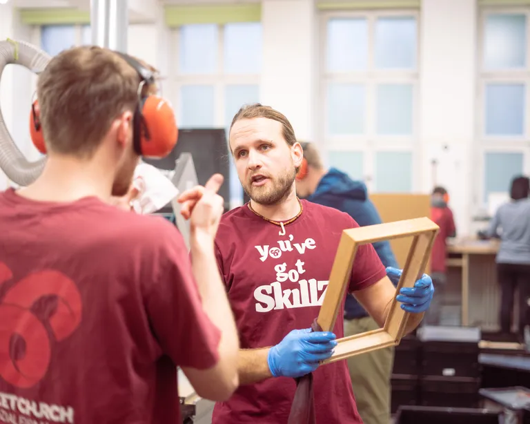 Zwei Männer unterhalten sich in einer Werkstatt; einer hält einen Holzrahmen, im Hintergrund arbeiten mehrere Personen.