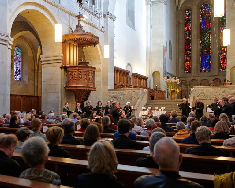 Viele Menschen sitzen in Kirchenbänken, vorne stehen Musiker und Sänger, Sonnenlicht fällt durch bunte Kirchenfenster.