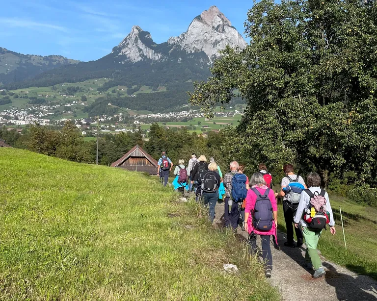 ​​Gruppe von 12 Seniorinnen und Senioren auf Wanderweg mit Rucksäcken, Blick auf die Mythen und Hügellandschaft bei Schwyz unter blauem Himmel​.