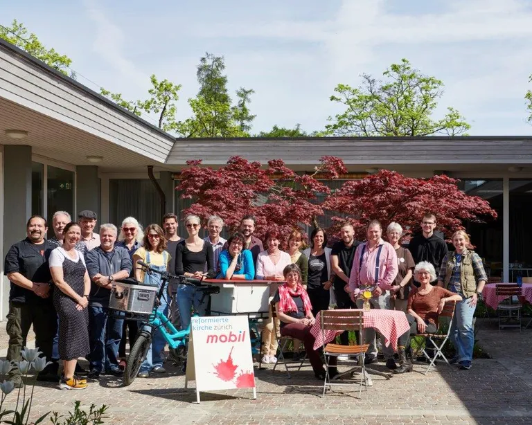 Eine Gruppe von Menschen steht und sitzt vor einem Gebäude neben einem roten Ahornbaum und einem Tisch mit Kuchen und Getränken.