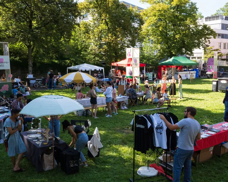 Menschen schlendern über einen Flohmarkt im Freien mit Ständen, Kleidung und Sonnenschirmen auf einer grünen Wiese.