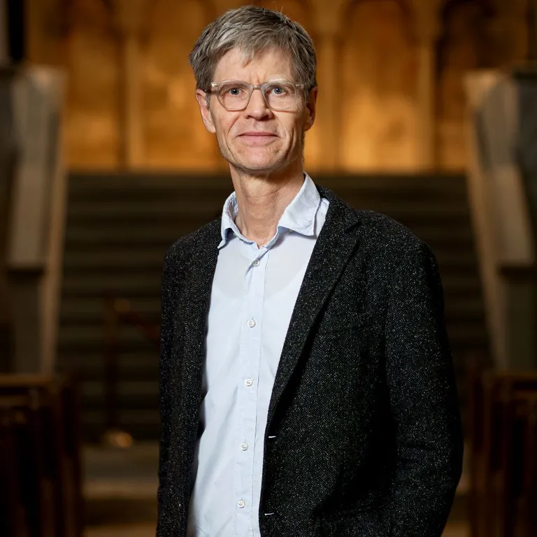 Pfarrer Martin Rüsch steht in der Kirche Grossmünster vor Treppen und bunten Kirchenfenstern, trägt Sakko und Hemd.