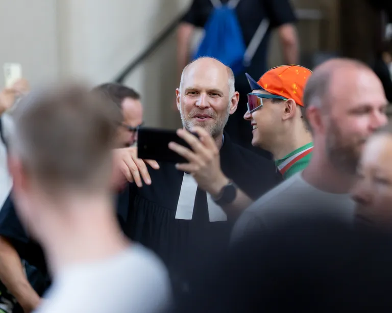 Mehrere Menschen stehen zusammen; ein Mann in der Mitte hält ein Smartphone und macht ein Selfie, die Szene ist unscharf im Vordergrund.