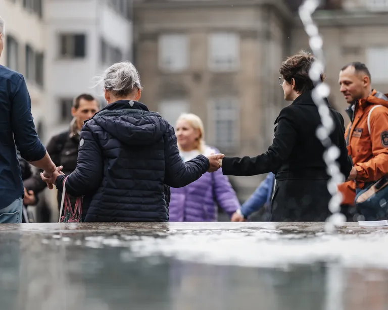 Mehrere Erwachsene stehen im Kreis und halten sich an den Händen, im Vordergrund ist ein Springbrunnen zu sehen.