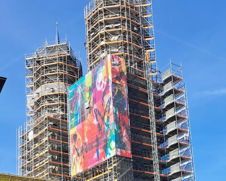Hohe Gebäude im Baugerüst, bunte Kunstbanner an den Fassaden, blauer Himmel im Hintergrund.
