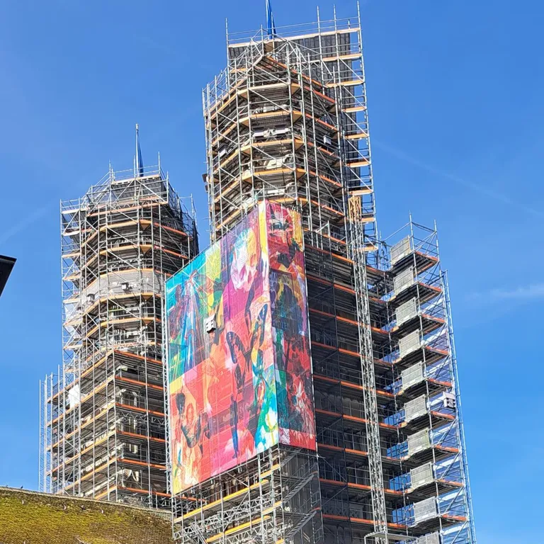 Hohe Gebäude im Baugerüst, bunte Kunstbanner an den Fassaden, blauer Himmel im Hintergrund.