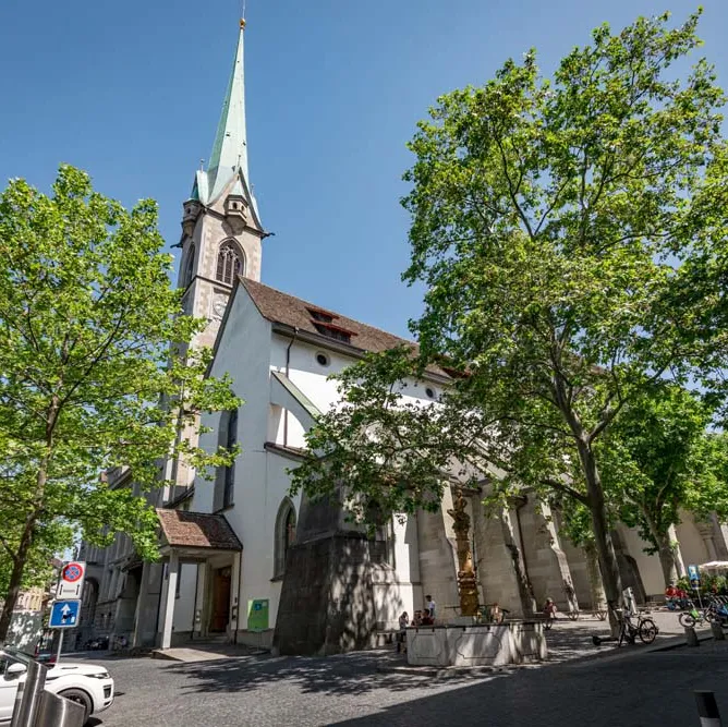 Alt-Text: Kirche mit hohem Turm an einer Straße, umgeben von grünen Bäumen und Autos bei sonnigem Wetter.