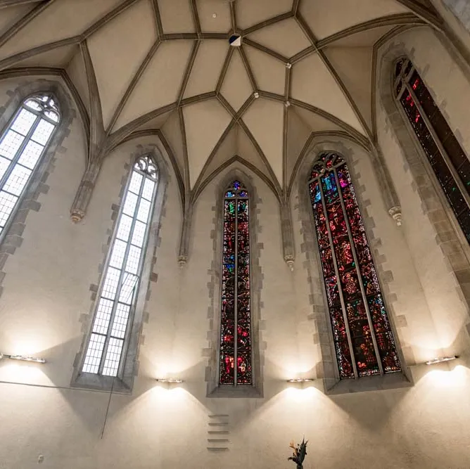 Hohes Kirchengewölbe mit fünf hohen, bunten und klaren Fenstern, gotischer Stil, beleuchtet von Wandlampen.