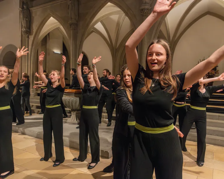 Mehrere Frauen tanzen synchron mit erhobenen Armen in einer Kirche oder Kathedrale. Sie tragen schwarze Kleidung mit gelben Gürteln.