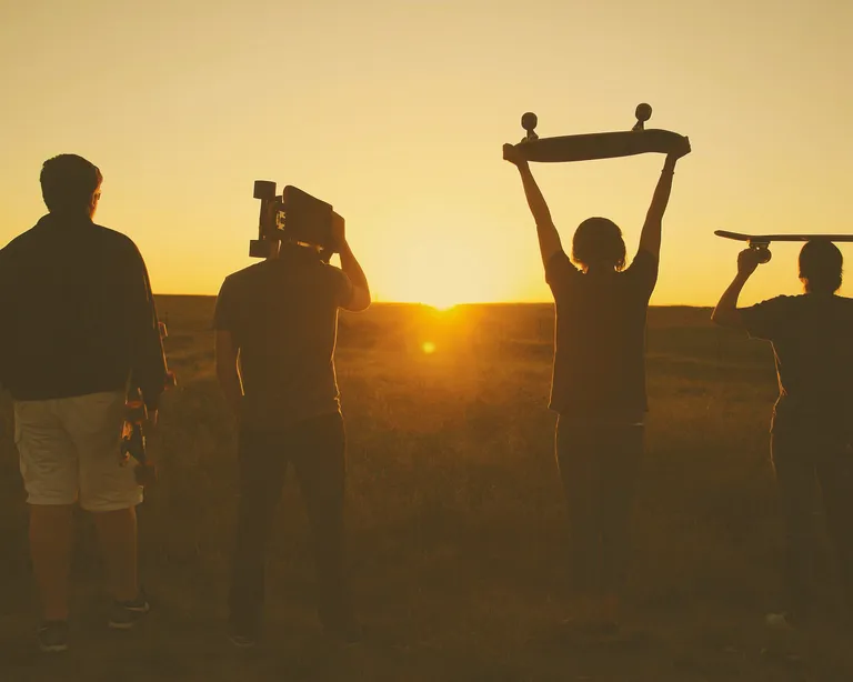 Vier Personen stehen bei Sonnenuntergang im Freien, zwei halten Skateboards in die Luft.