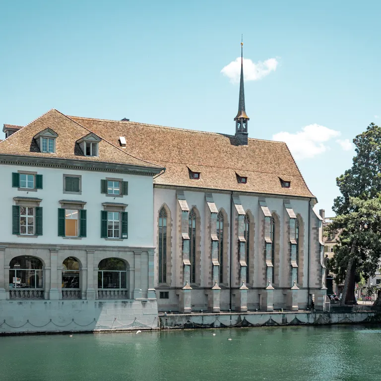 2 historisches Gebäude: links das Helmhaus, rechts die Wasserkirche mit spitzem Türmchen am Fluss, daneben Bäume, oben blauer Himmel.