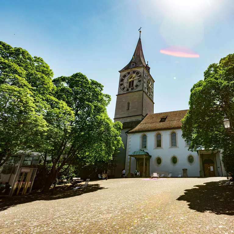 A historic church with a tower, surrounded by trees in a sunny square.