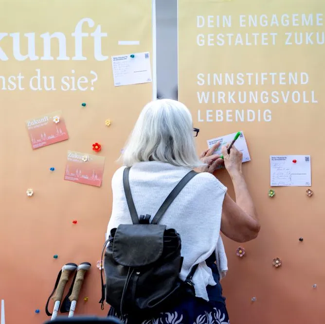Eine Person mit weißem Haar und Rucksack schreibt an eine orange Wand mit Zukunfts-Fragen und Karten.