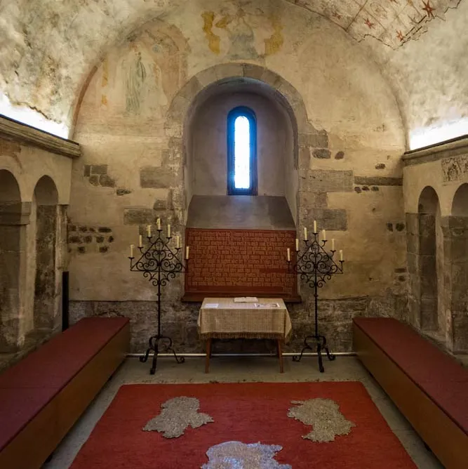 Steinerner Raum mit Gewölbedecke, roten Bänken, kleinem Altar und schmalem Fenster; alte, teils beschädigte Wand- und Deckenmalereien.