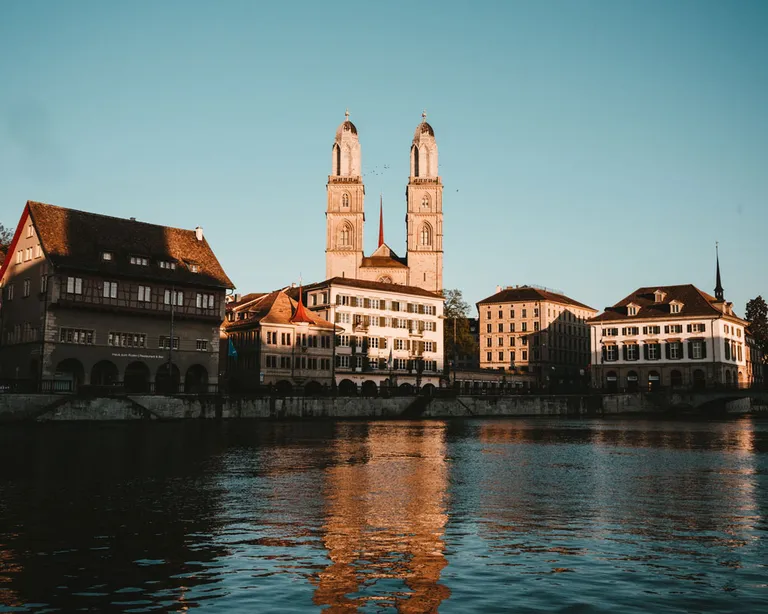 Alt-Text: Zwei Türme einer Kirche spiegeln sich im ruhigen Wasser, Altbauten am Ufer, blauer Himmel.