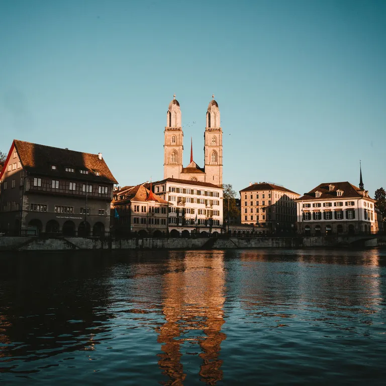 Alt-Text: Zwei Türme einer Kirche spiegeln sich im ruhigen Wasser, Altbauten am Ufer, blauer Himmel.