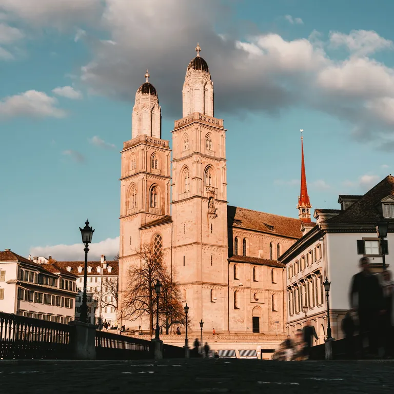 The Grossmünster with its two towers at sunset, surrounded by historic buildings and a blue sky with clouds.