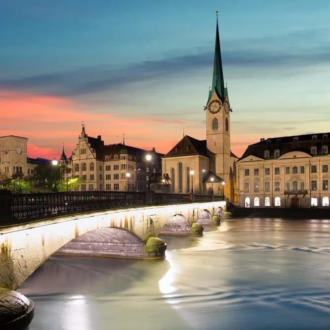 Abendliche Stadtansicht mit beleuchteter Brücke über einem Fluss und einer Kirche mit hohem Turm im Hintergrund.