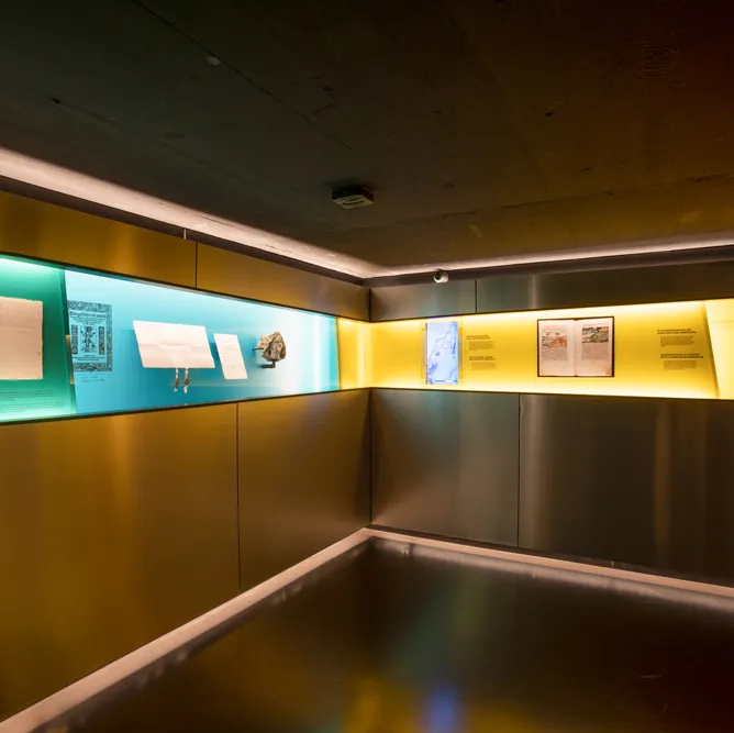 Ein moderner Ausstellungsraum mit beleuchteten Vitrinen an den Wänden, in denen Dokumente und Bilder gezeigt werden.