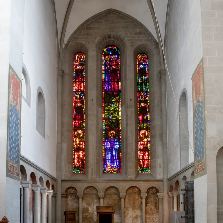 Hoher Kirchenraum mit buntem Glasfenster von Augusto Giacometti im Altarbereich, helle Wände, steinerner Boden und einige Fresken.