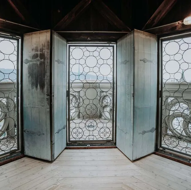 Blick aus einem Raum mit kunstvoll verzierten Fenstergittern und Tür ins Freie, helle Holzplanken am Boden.
