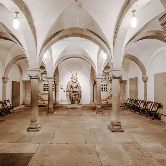 Gewölbte Krypta mit steinernen Säulen, Stühlen an den Wänden und einer religiösen Statue im Hintergrund.