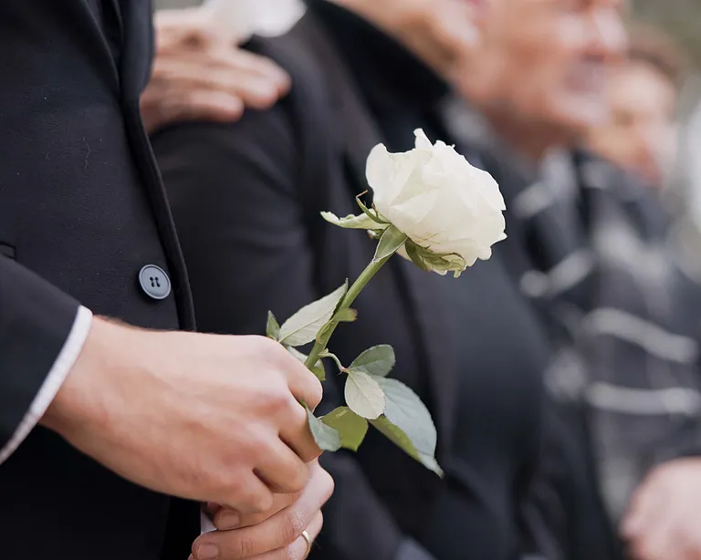 Mehrere Menschen in dunkler Kleidung, eine Person hält eine weiße Rose, unscharfer Hintergrund, traurige Stimmung.