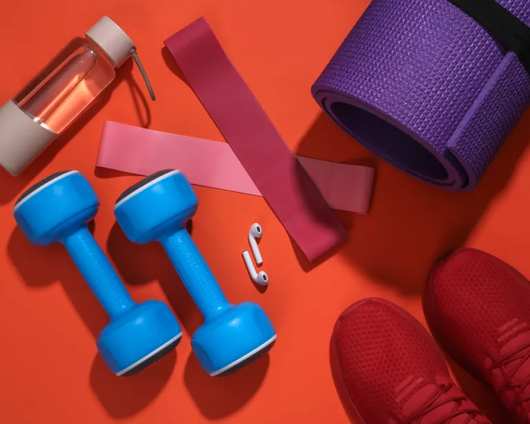 Fitnessausrüstung auf orangem Hintergrund: blaue Hanteln, Trinkflasche, Gymnastikmatte, Widerstandsbänder, Kopfhörer und rote Sportschuhe.