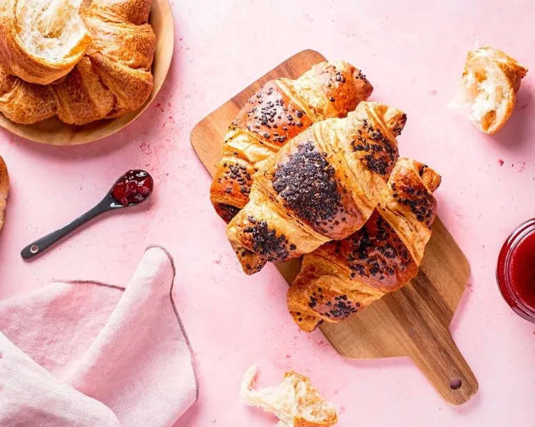 Drei Croissants auf einem Holzbrett, daneben Marmelade, ein Löffel und weitere Backwaren auf rosa Untergrund.