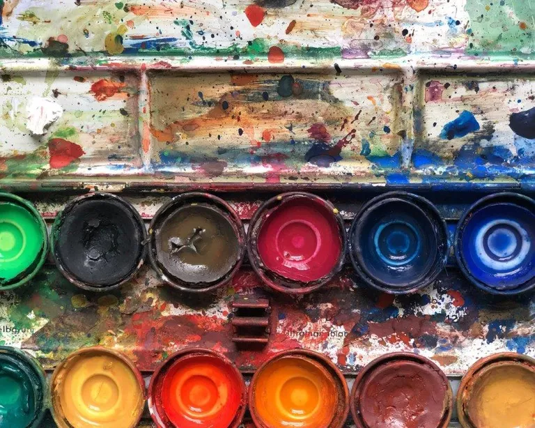 Offene Aquarellfarbpalette mit mehreren bunten, teils benutzten Farbnäpfen und Farbspritzern auf dem Deckel.