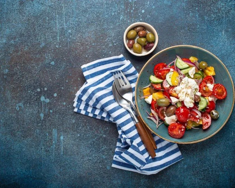 Bunter griechischer Salat mit Feta, Tomaten, Gurken und Oliven auf einem Teller, daneben eine Schale Oliven und Besteck auf Stoffserviette.