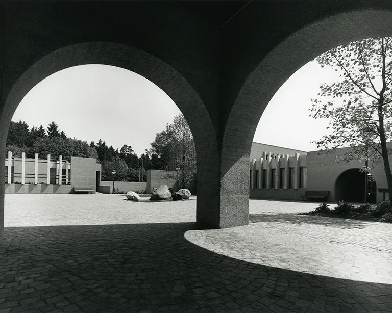 Schwarzweißfoto von zwei großen Rundbögen, im Hintergrund moderne Gebäude, Bäume und geparkte Autos.