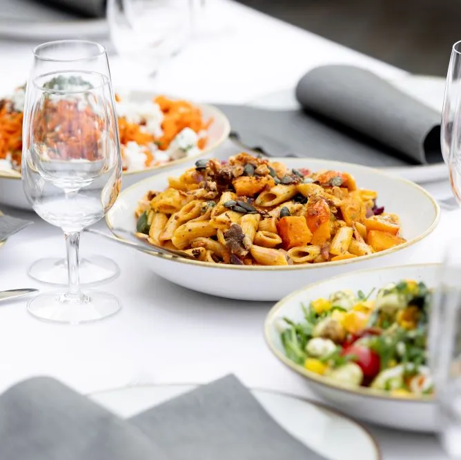 Weiße Tafel mit Tellern Pasta, Salat und Fisch, graue Servietten, Weingläser und Besteck, festlich gedeckt.