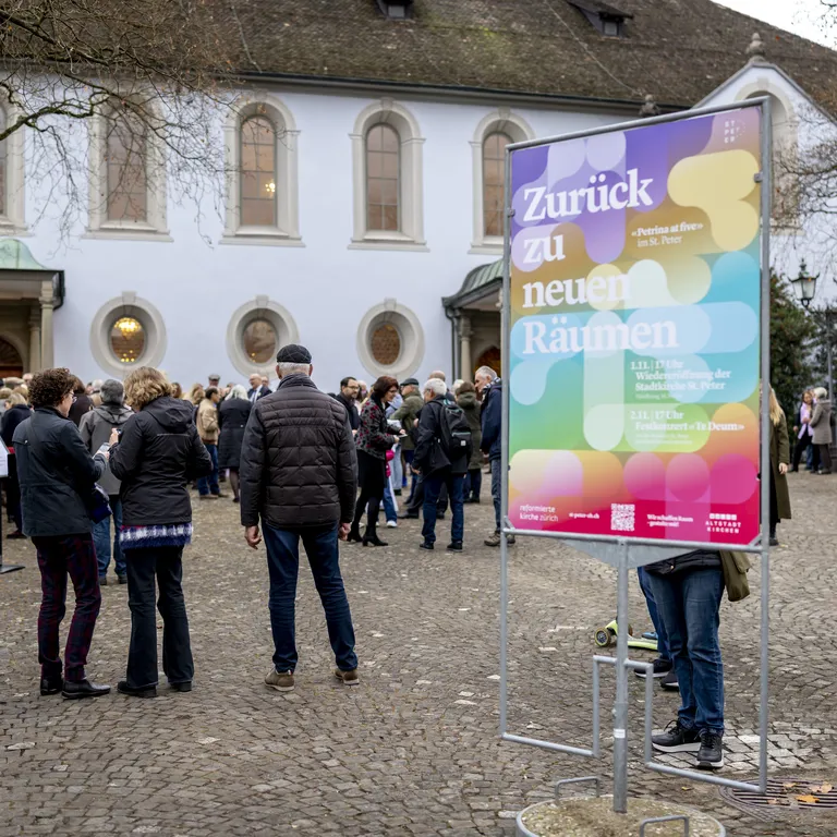 Eine Menschenmenge steht vordem St. Peter in Zürich, im Vordergrund sieht man ein buntes Plakat auf einem Ständer.