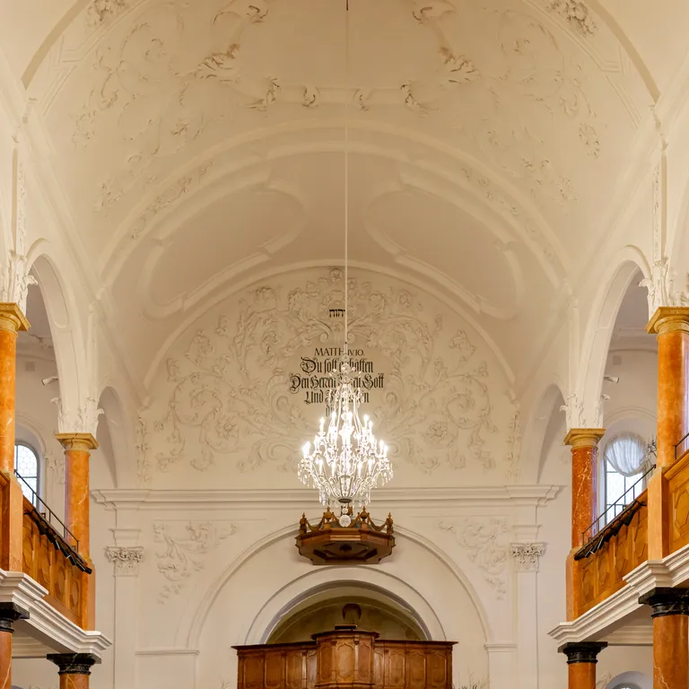 Heller barocker Kircheninnenraum mit hohen Gewölbedecken, Kronleuchter und Emporen aus Holz auf beiden Seiten.