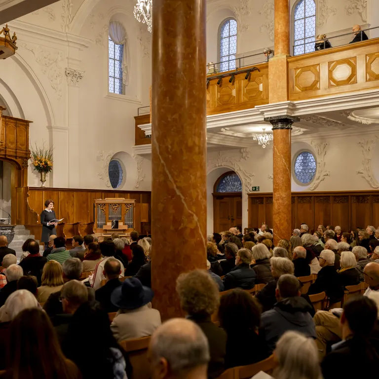 Innenraum der Kirche Grossmünster mit Kanzel, Taufbecken, Bänken und bunten Glasfenstern am Altar, stimmungsvolles Licht.
