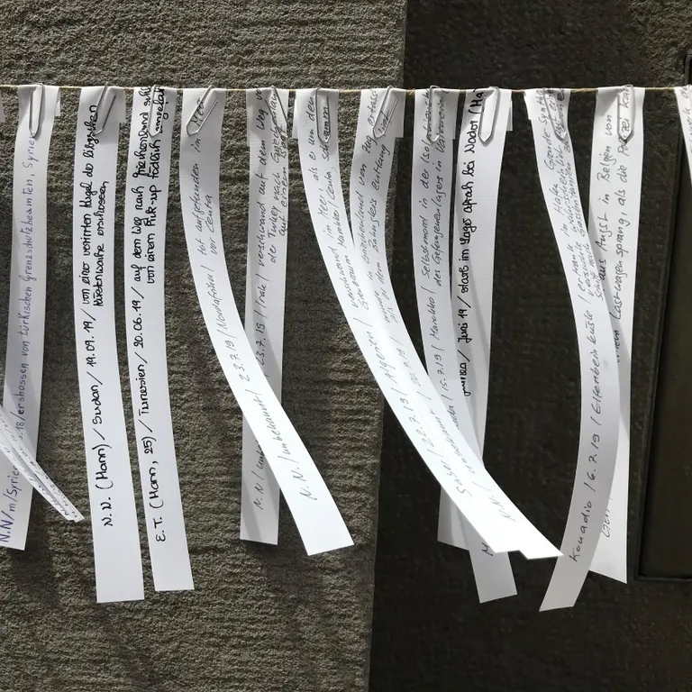 Mehrere bedruckte Papierstreifen mit Namen hängen nebeneinander an einer Leine vor einer grauen Wand.