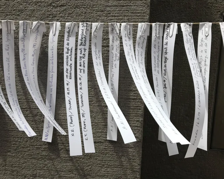 Mehrere bedruckte Papierstreifen mit Namen hängen nebeneinander an einer Leine vor einer grauen Wand.