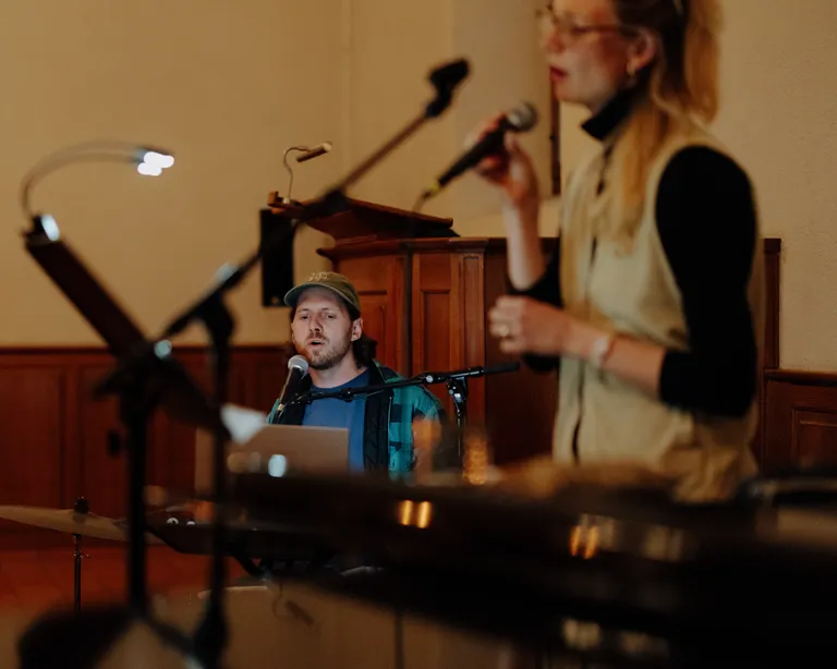 Zwei Personen singen in einem Saal ins Mikrofon, eine sitzt am Schlagzeug, die andere steht im Vordergrund.