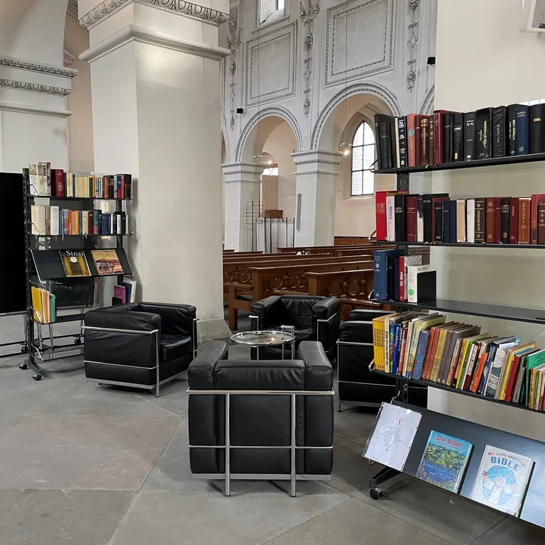 Regale mit Büchern und schwarze Sessel stehen in einer hellen Kirchenhalle mit hohen Bögen und großen Fenstern.