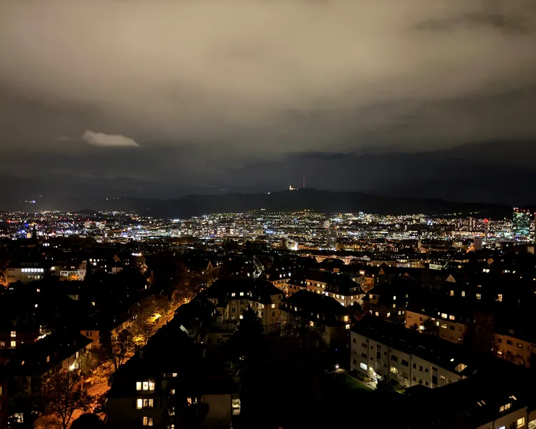 Blick auf die Stadt Zürich bei Nacht mit vielen leuchtenden Lichtern und bewölktem Himmel im Hintergrund.