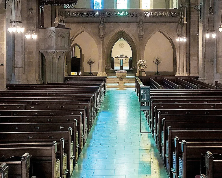 Innenraum einer Kirche mit Steinbögen, leeren Holzbänken und bunten Fenstern am Altar.